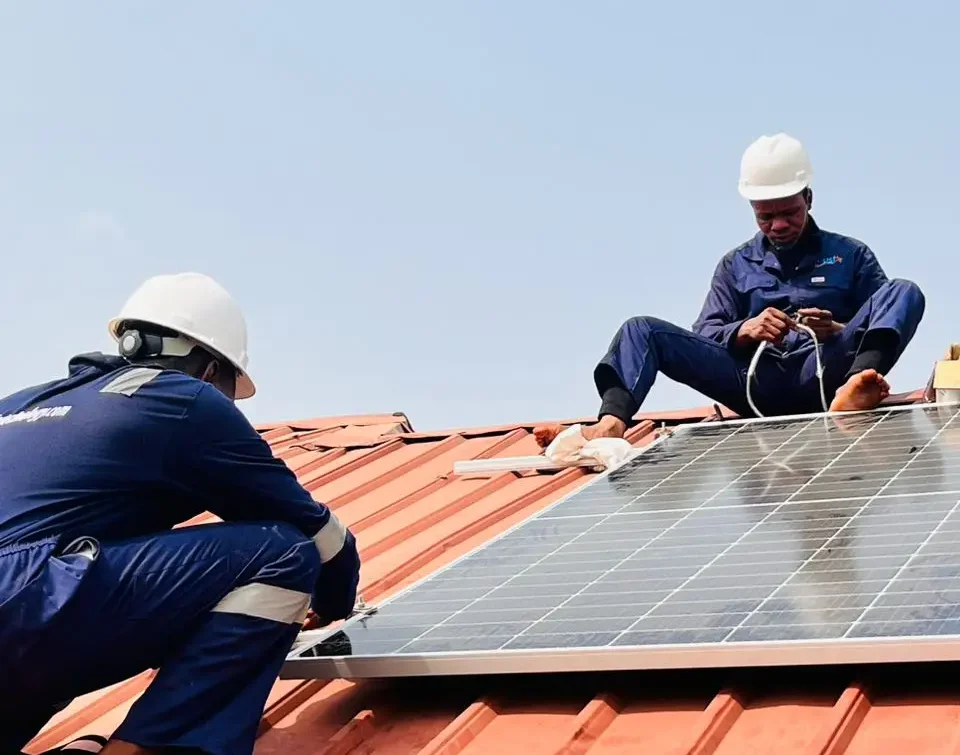 Two Corustar technicians in full safety gear mounting a solar panel on a zinc roof, with one connecting cables and the other securing the panel