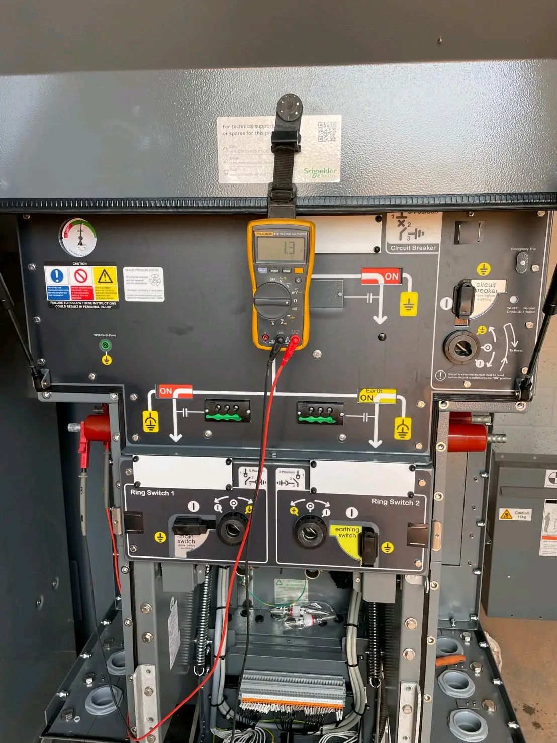 Open electrical panel with labeled components and a connected multimeter displaying test readings.